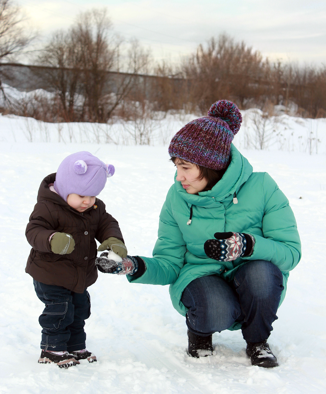 bundled up mom and toddler make a snowball in a snowy field