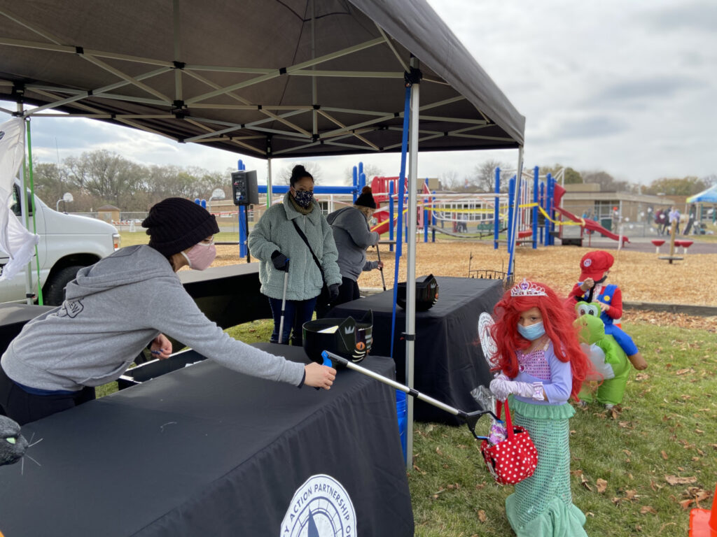 masked CAP-HC staff person uses arm-extending tool to give candy to a masked girl dressed as a mermaid at the 2020 Spooktacular